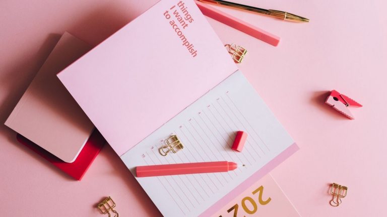 Organized workspace with pink planner, pen, and clips on pastel background.